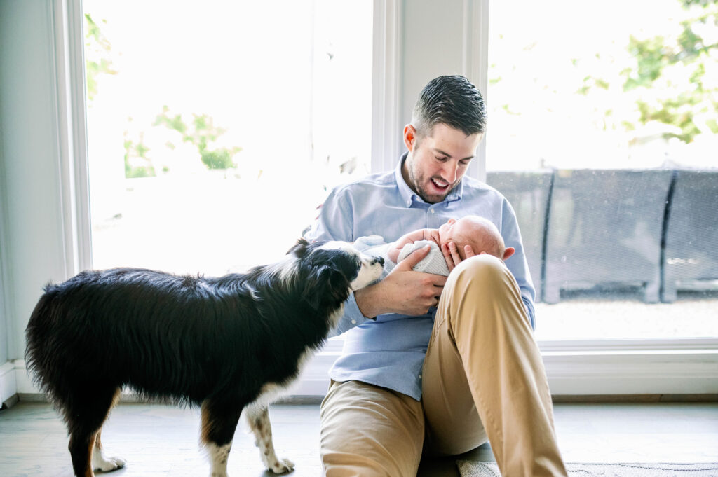 dad smiles at newborn while dog watches