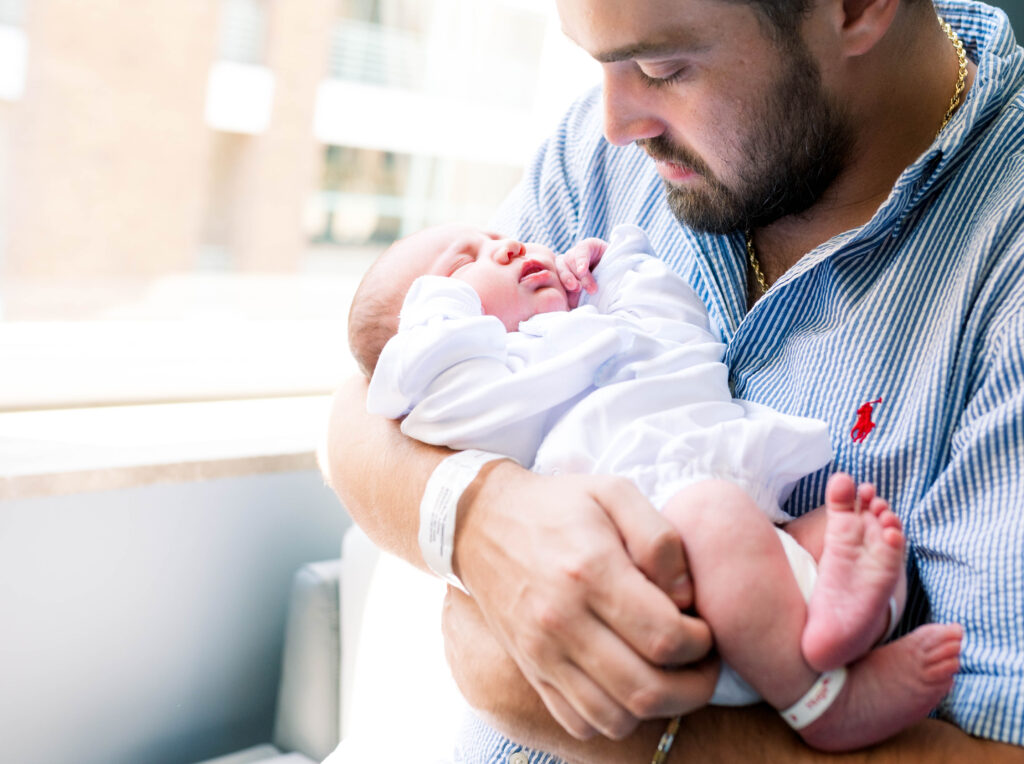 Dad is holding his newborn son near a window in the hospital room for a fresh 48 session.
