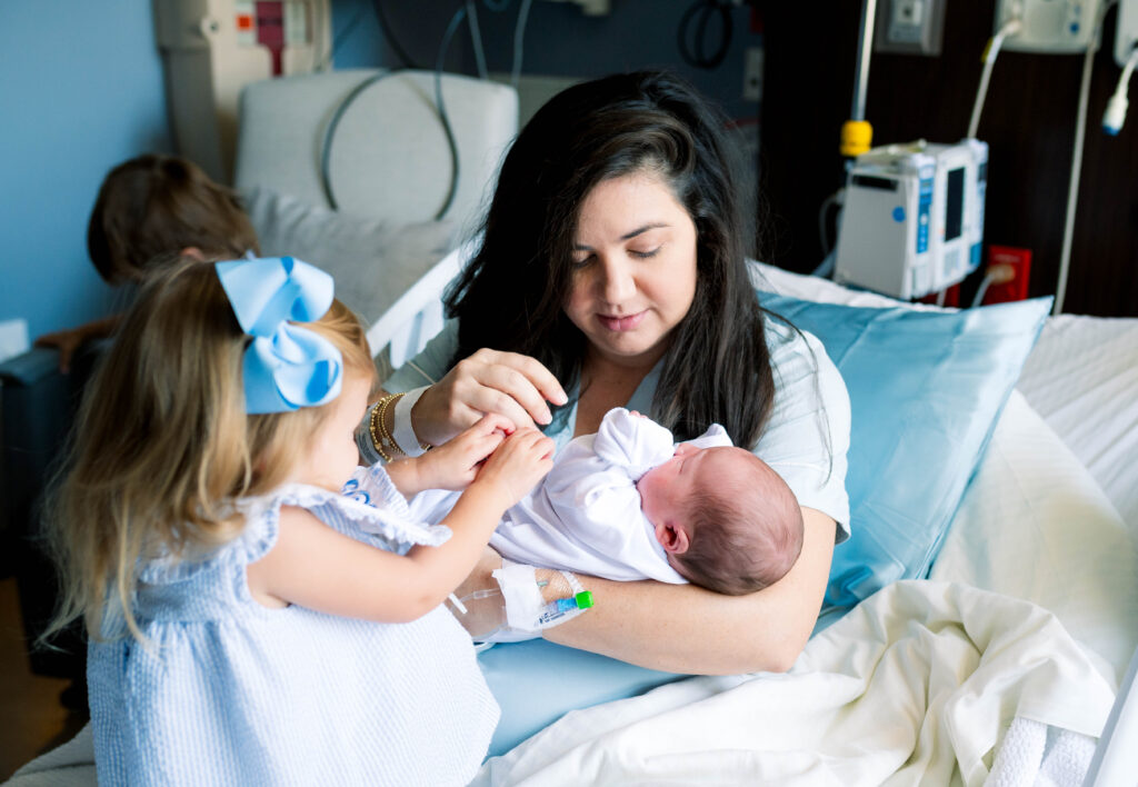 A mom in a hospital bed is holding her newborn son while her daughter is helping comfort him.

