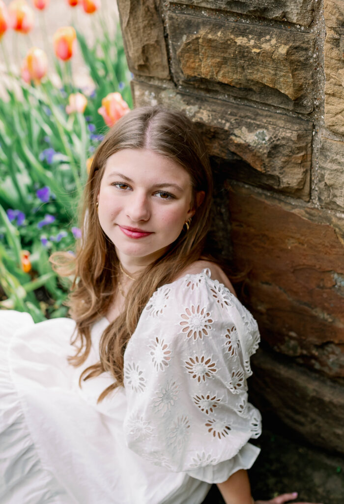A teenage girl sits against a stone wall during her fall senior photos.
