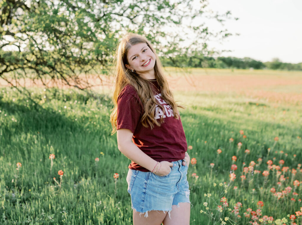 A senior teenager in a field with wildflowers is wearing her college shirt during her fall senior photo session.
