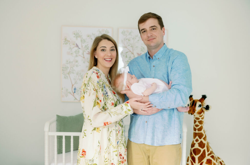 Mom and dad proudly hold newborn girl while standing in a beautiful nursery.
