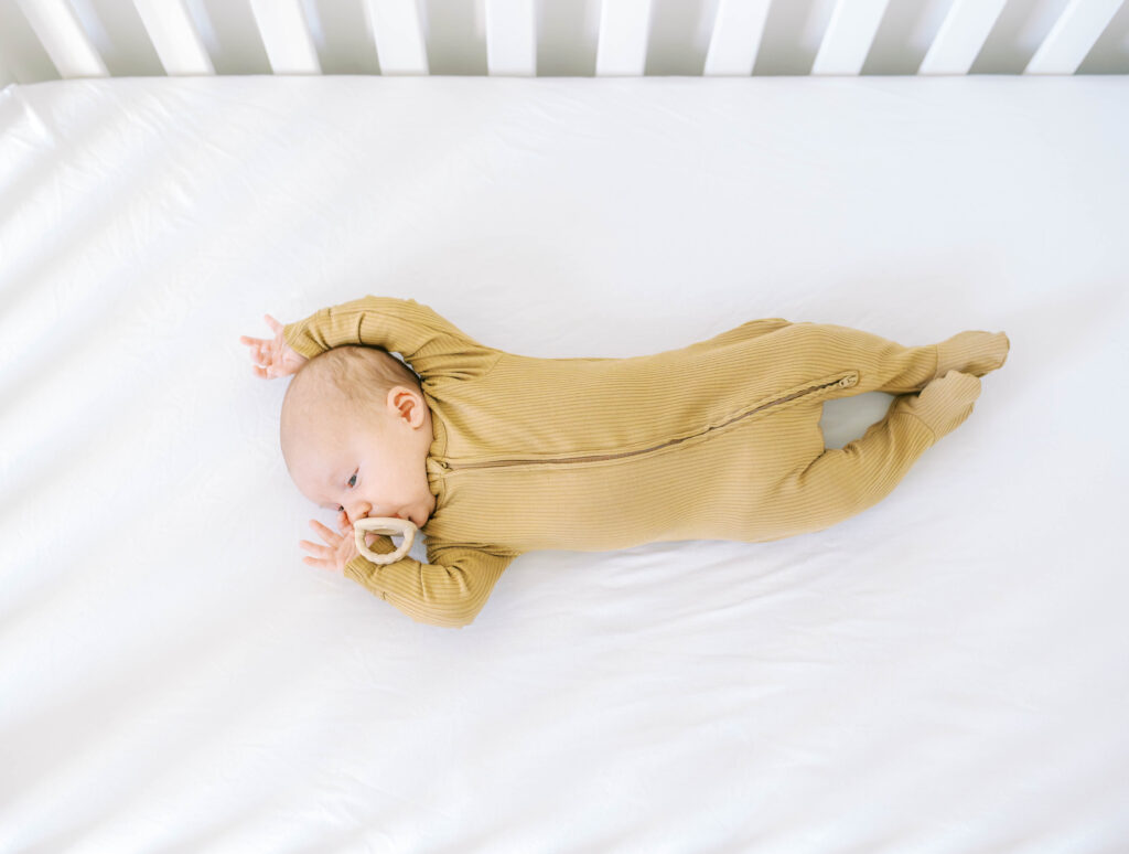 Newborn wearing a close fitting onesie stretches out in crib during photo session with Aledo photographer Allison Krogness.