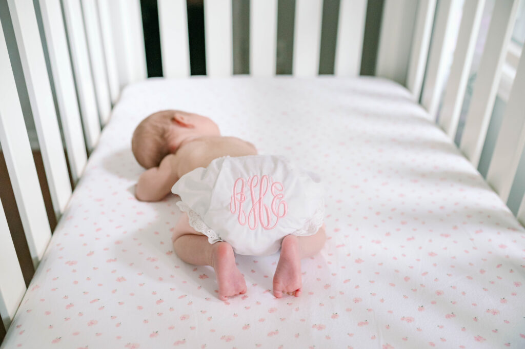 A baby girl is sleeping in her crib, wearing a pink monogrammed diaper cover.