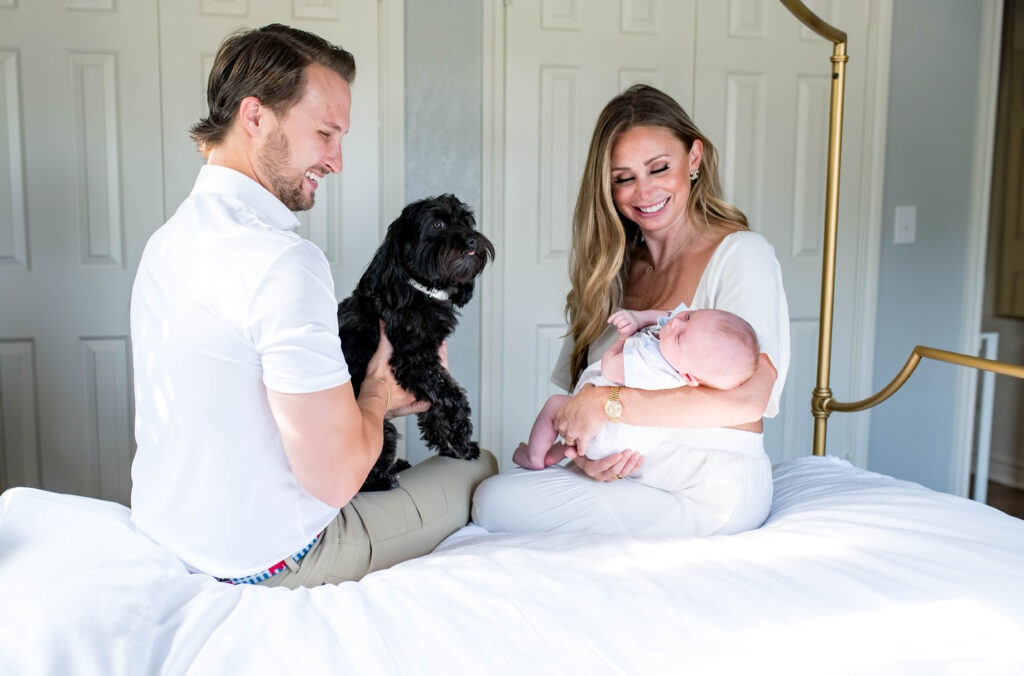 Parents are holding their newborn son on the bed while the family dog looks on.
