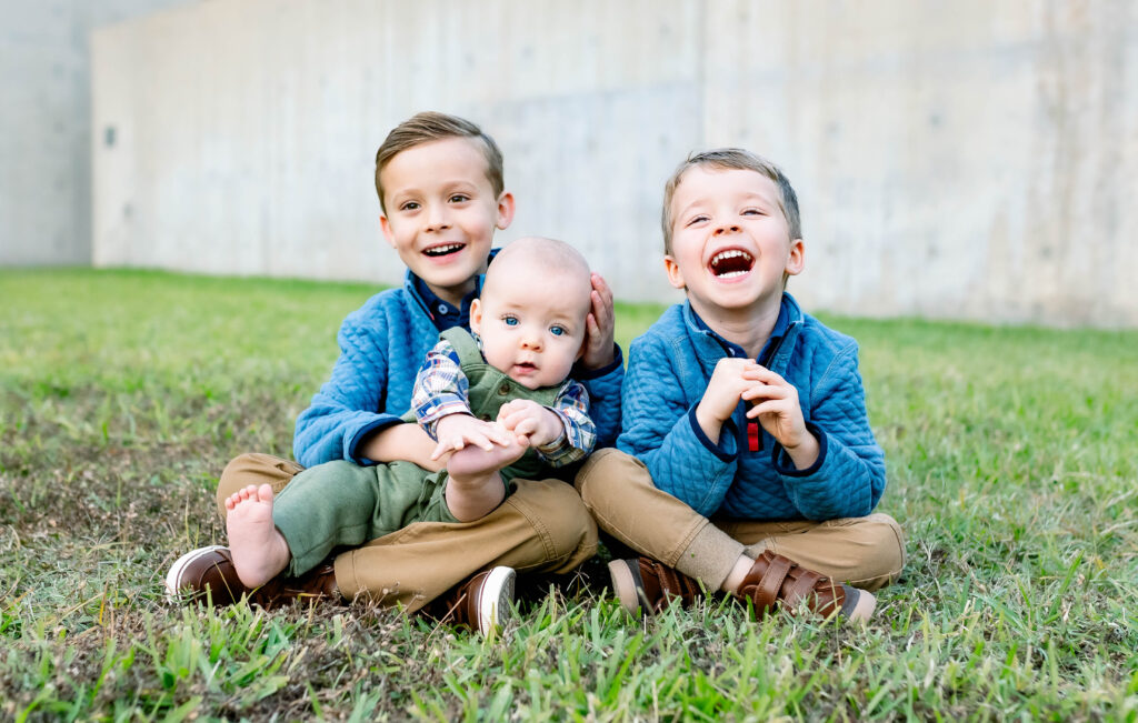 Three young brothers sitting in the grass smiling at the camera during outdoor fall mini session.
