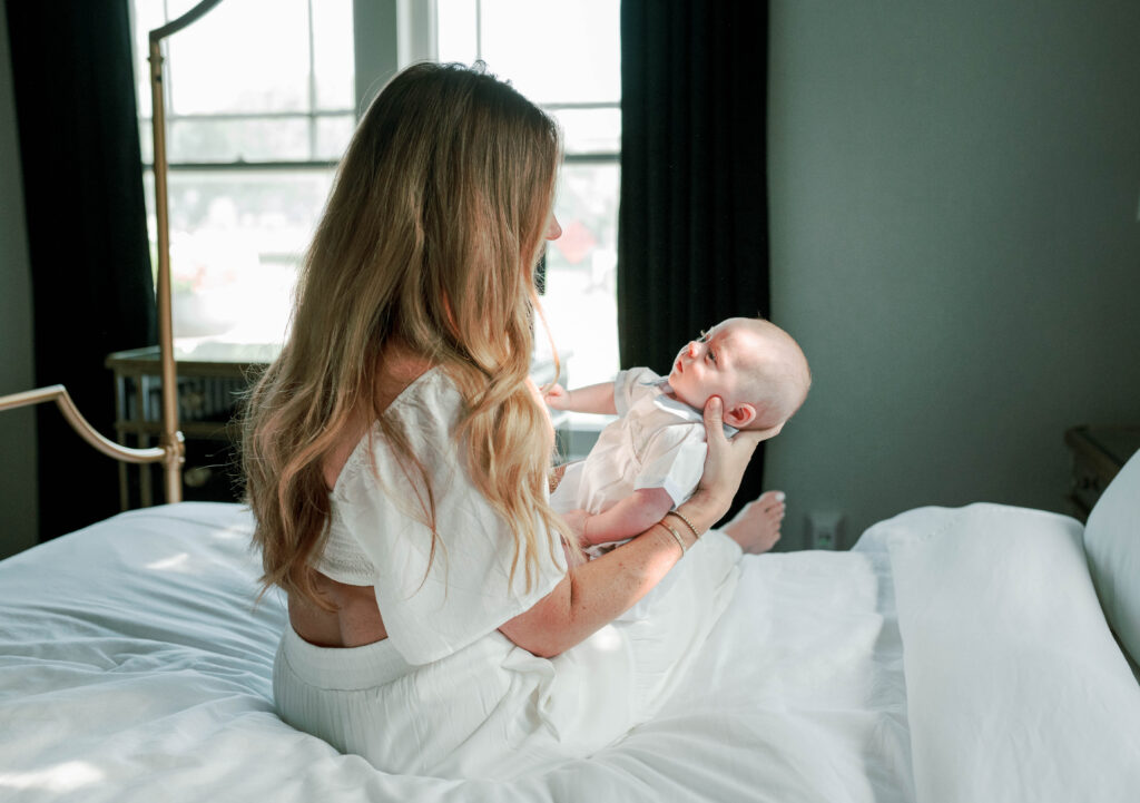 Mother sitting on the bed, cradling newborn son during an in-home photo session with Allison Krogness.

