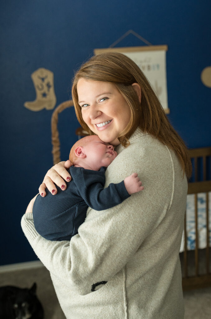 A new mom is standing in the nursery holding her newborn son, looking at the camera during a newborn session with Allison Krogness.