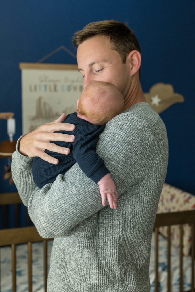 A new dad is holding his newborn son in the nursery with his hand on his baby's back.