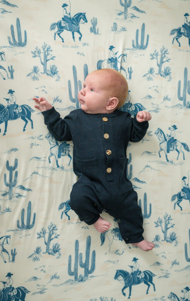 A newborn boy is lying in his crib on cowboy-themed sheets, during a lifestyle newborn session in Arlington TX.