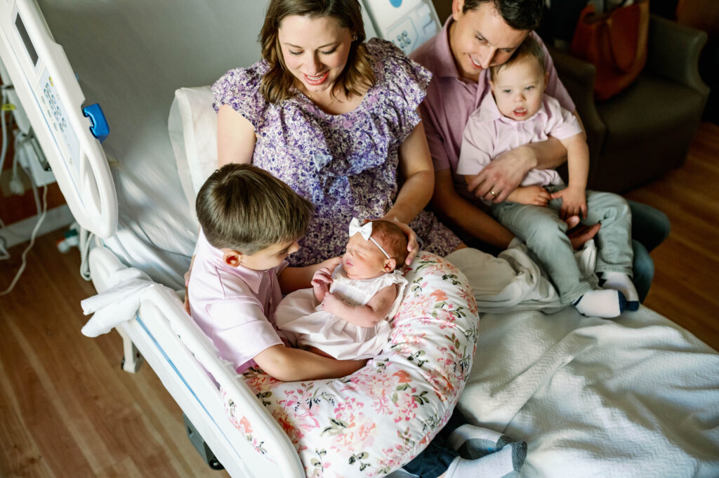 The oldest brother is holding his newborn baby sister while sitting on his mom's hospital bed.