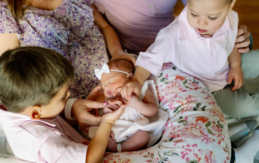Two older brothers are lovingly looking at and holding hands with their newborn sister during a Fresh 48 Session.

