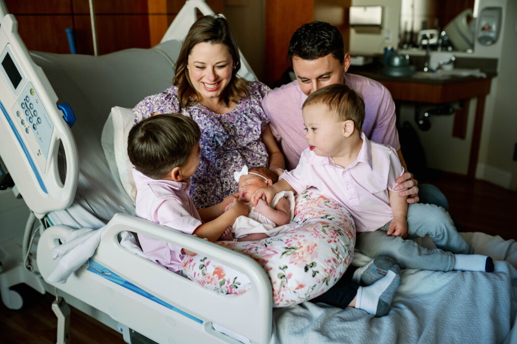 Mom and Dad are smiling at their two older children, who are holding their newborn sister in the hospital room.