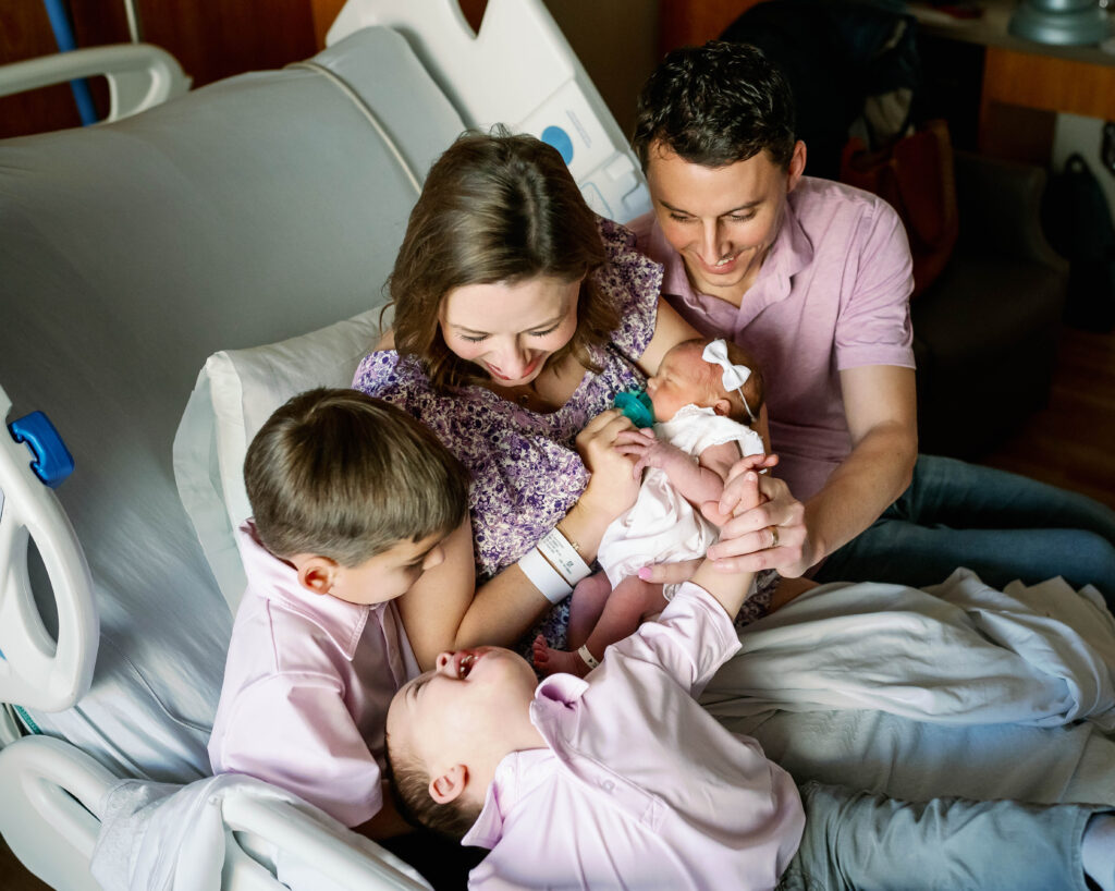 Mom, Dad, and two older siblings are helping to hold their newborn sister while in the hospital.
