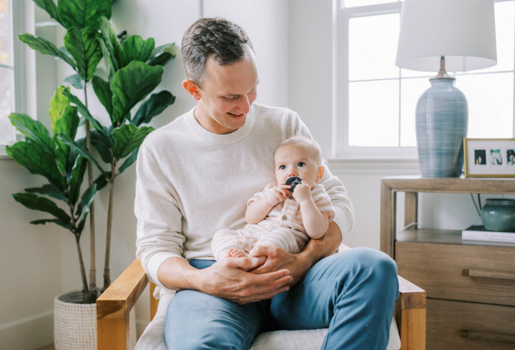 Fort Worth newborn photographer, Allison Krogness, captures a dad holding a newborn in a chair near a window.
