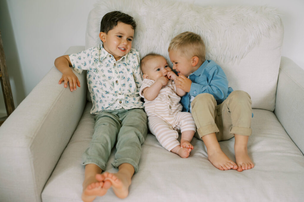 Two older brothers carefully hold their newborn brother while sitting together in a large soft chair.

