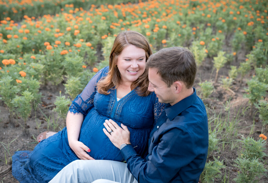 A couple sits in a field of flowers looking at each other during a maternity photo session.
