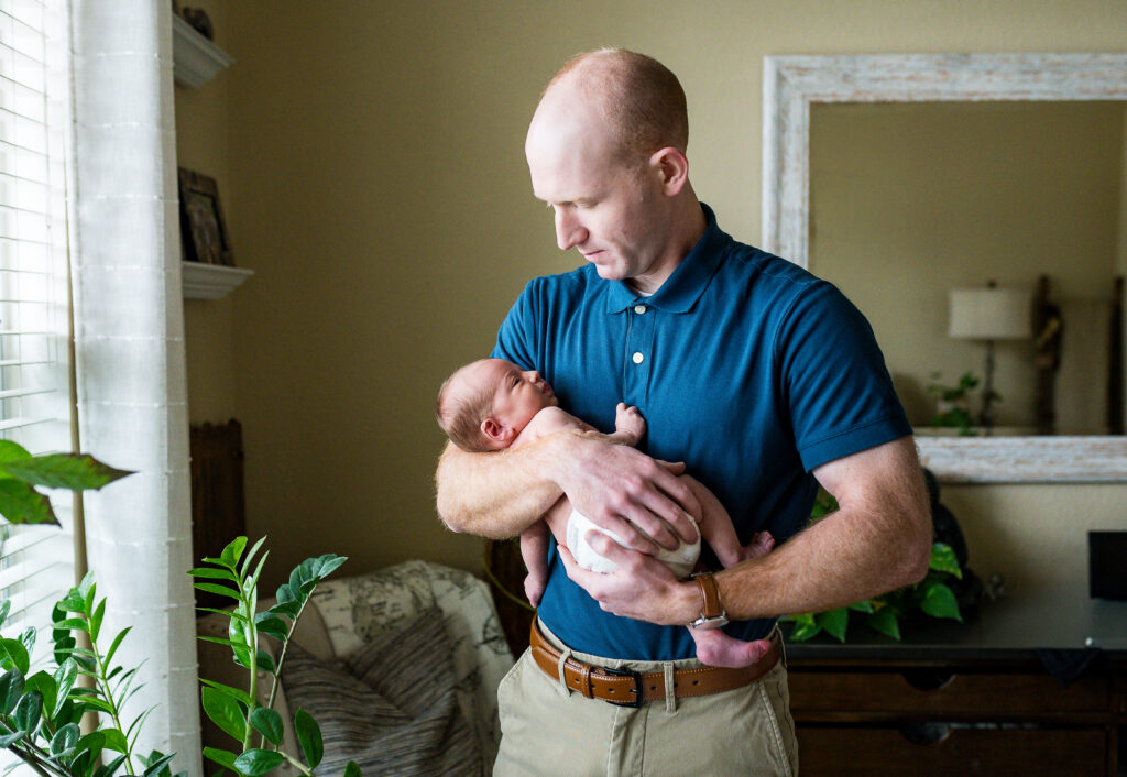 Dad is cradling his newborn son near a window during an in-home newborn session with Allison Krogness Photography.