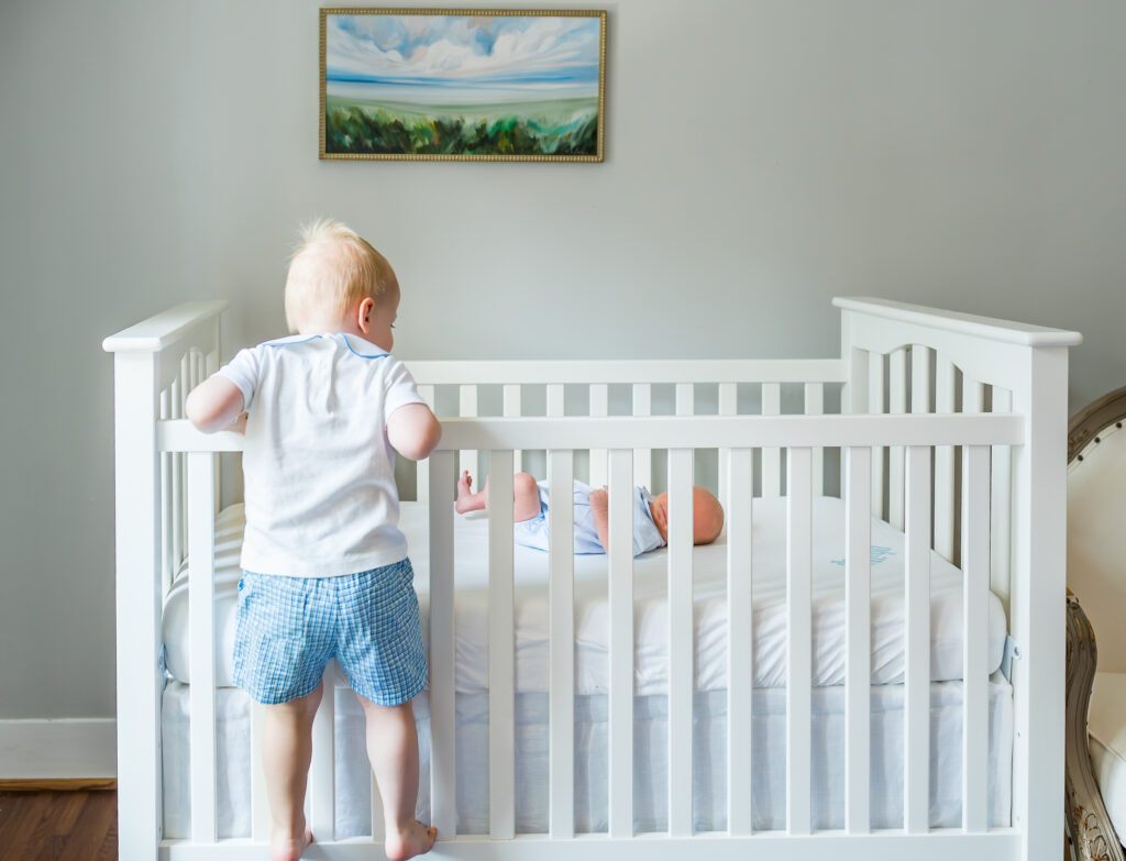 Older brother standing on crib edge looking down at his newborn brother in a beautiful, well-lit nursery.
