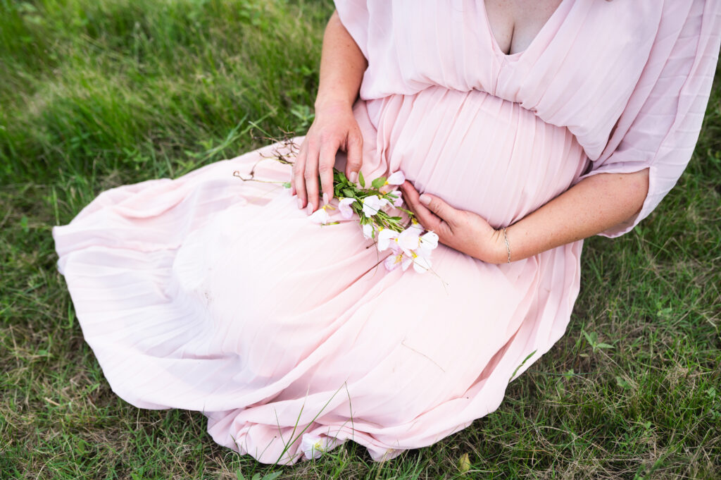 DFW maternity photographer Allison Krogness captures a mother sitting in a field with flowers wearing a pink dress.
