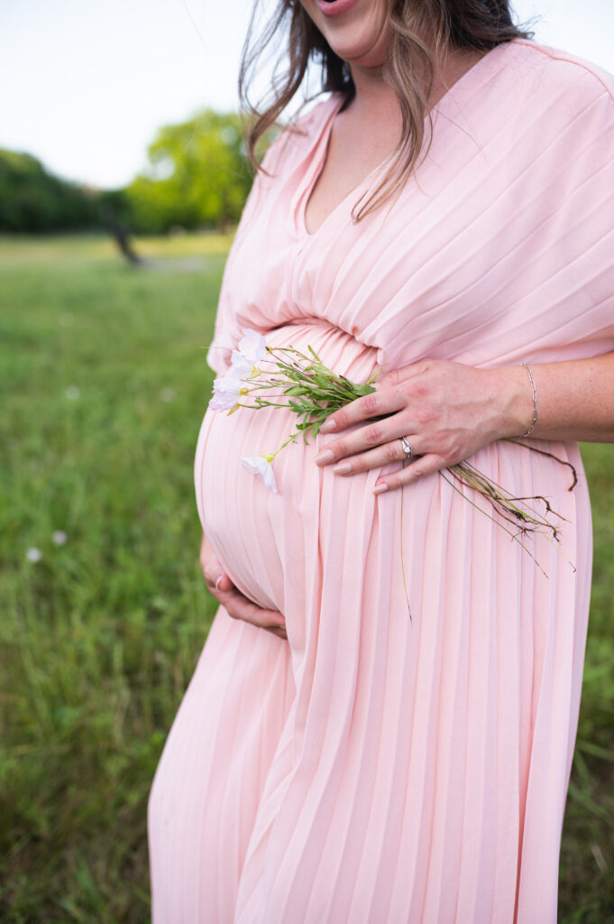 An expectant mother wearing a pink dress, holding flowers against her belly during an outdoor photo session.
