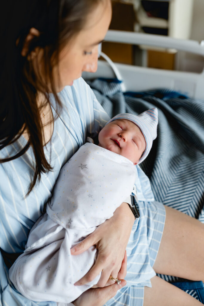 Fort Worth fresh 48 photographer, Allison Krogness captures a mom holding her newborn in a swaddle and cap sitting on the edge of a hospital bed.
