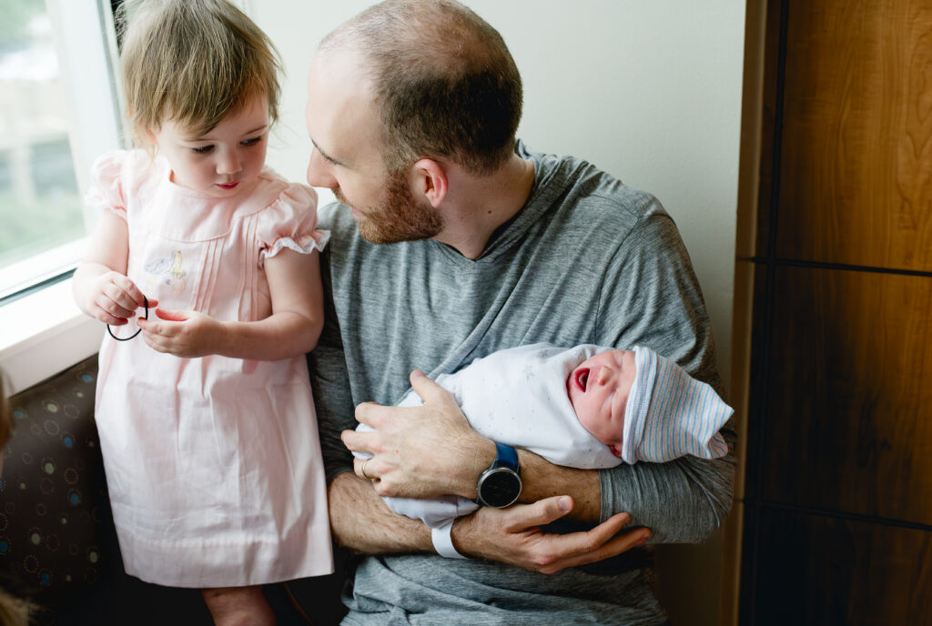 A dad holds his newborn while sitting next to his daughter and they talk about their new baby while looking out a hospital window.
