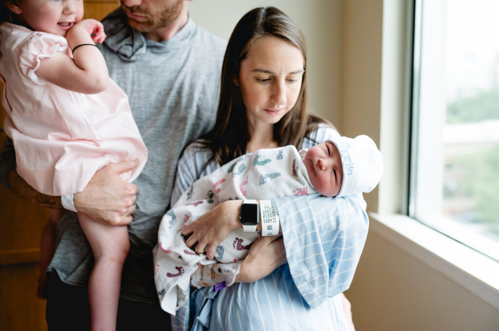 A newborn baby looks towards the camera while mom is holding the baby with dad and older sibling in the background.
