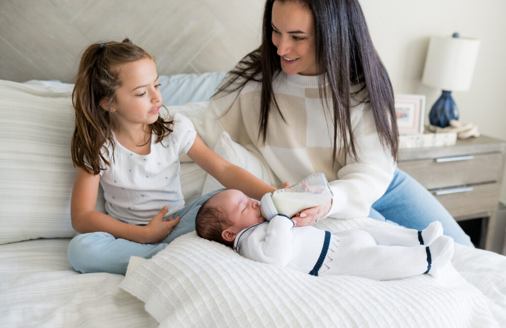 An older sister helps mom feed a bottle to her newborn brother while sitting on a bed.
