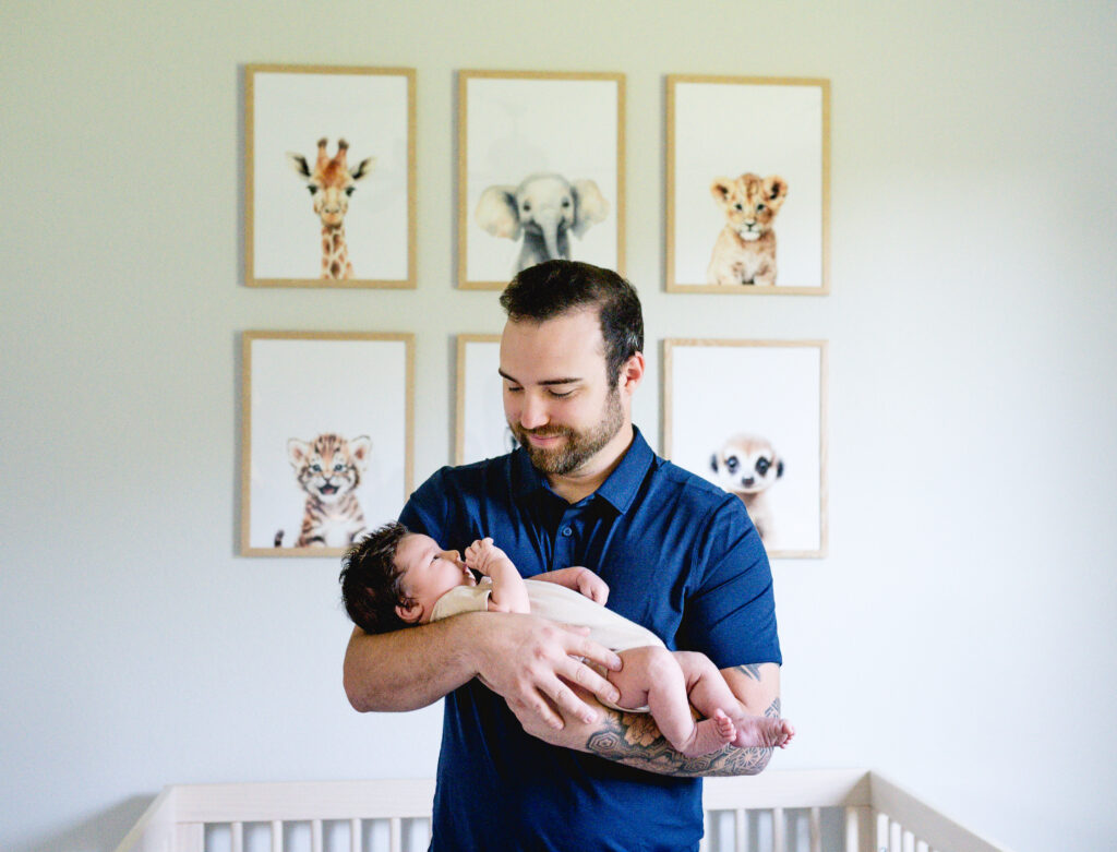 Dad holds newborn son in his nursery with animal prints above the crib
