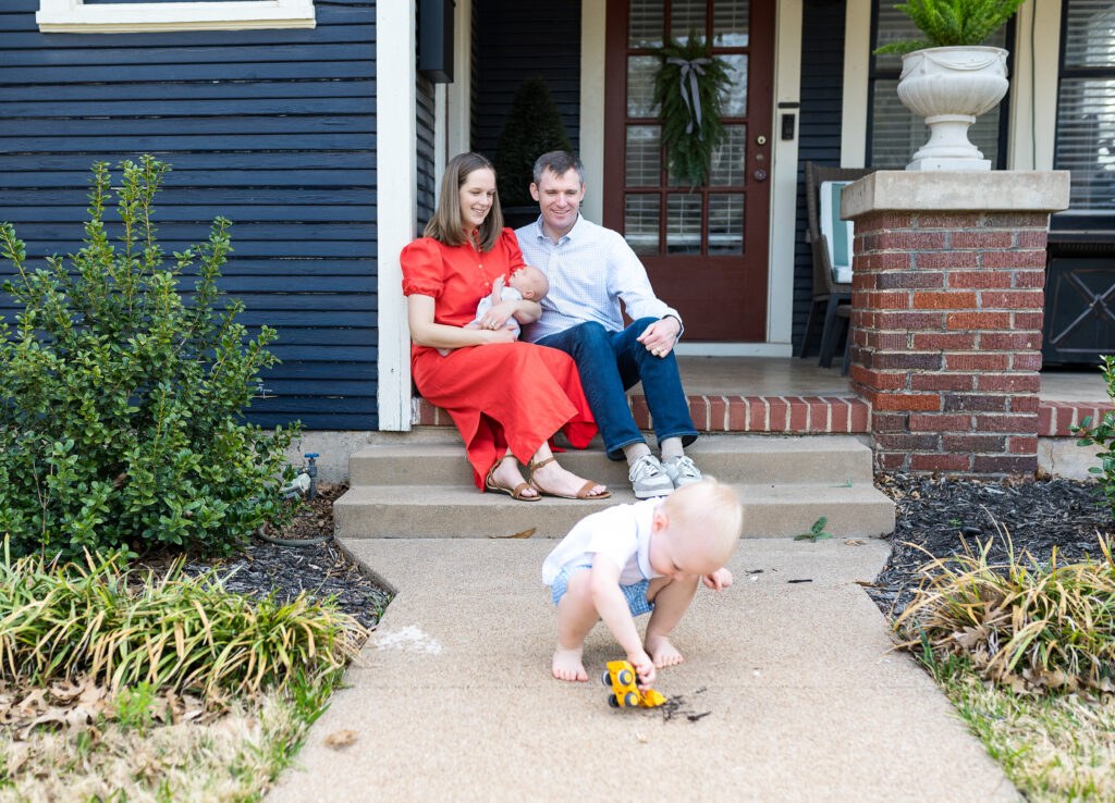 Mom and Dad holding a newborn baby together during a newborn photography session while their older child plays on the sidewalk.
