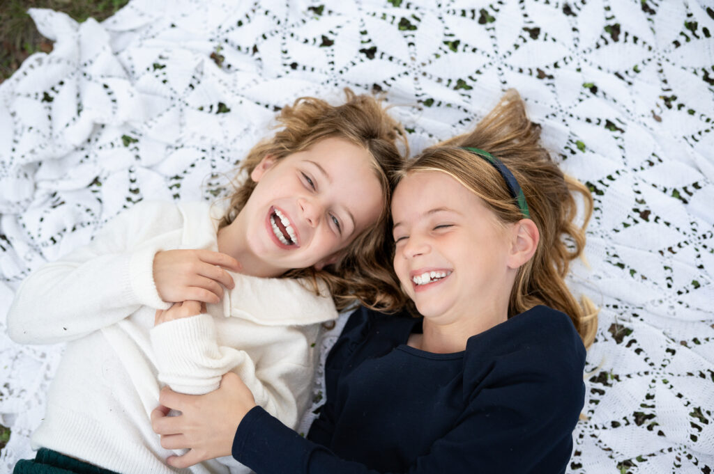 Two sisters lay on a vintage quilt laughing at the camera.
