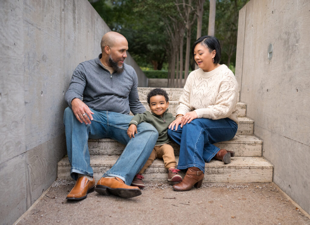 A mom and dad sit with their toddler between them on the steps of the Kimbell Art Museum for a Fort Worth fall mini-session photo shoot.
