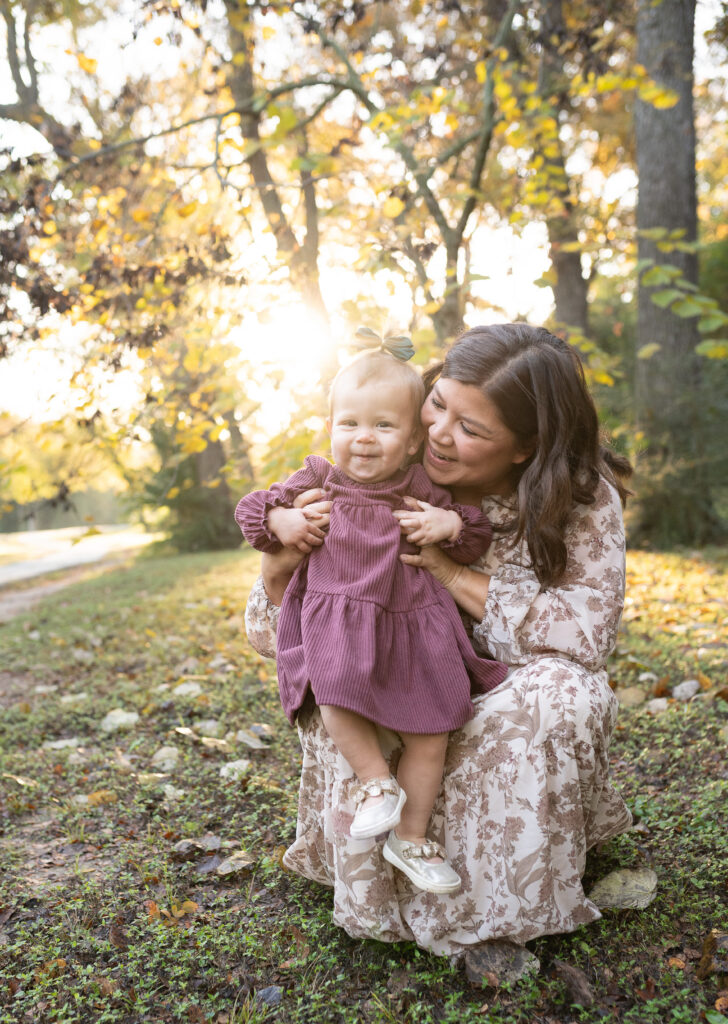 A mom snuggles with her toddler-aged daughter with beautiful light filtered behind them in the fall foliage.

