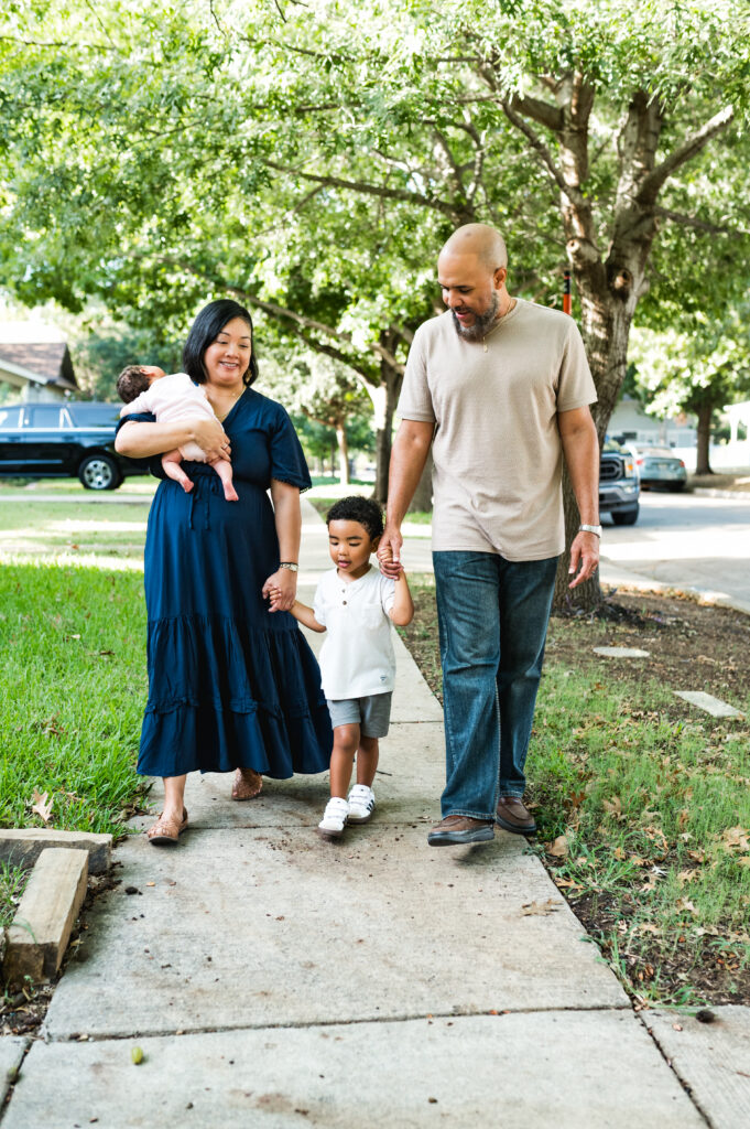 A family is walking together in their neighborhood, holding their newborn and toddler's hands.