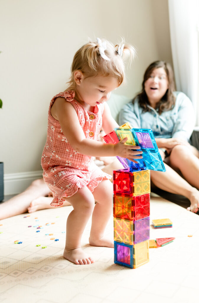 A toddler girl is building a tower while her family watches her during an in-home photo session.
