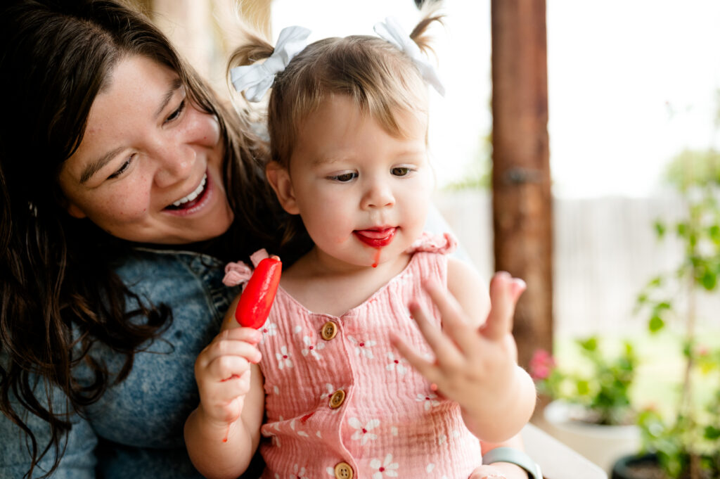 Fort Worth candid family photographer Allison Krogness captures a girl sitting on her mom's lap, enjoying a messy popsicle.

