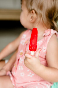 A toddler girl sits on her mom's lap, holding a red popsicle during a candid family photo session.
